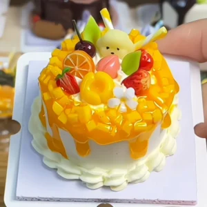 Finished example of a DIY fruit cake craft kit with yellow glaze-style topping, white cream-like trim, fruit accents, floral details, and decorative toppers
